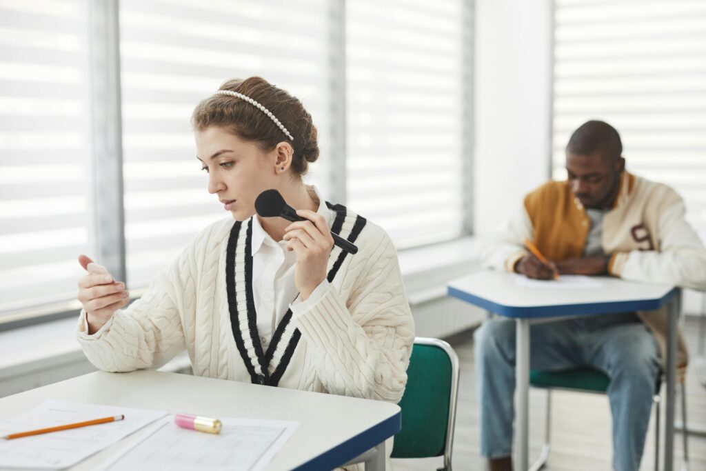 Two diverse students in a classroom, one using makeup, the other taking notes.